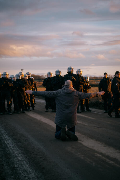 Ein Mann, von hinten fotografiert, der auf Knien und mit ausgebreiteten Armen einer Gruppe von Polizist:innen gegenübersteht